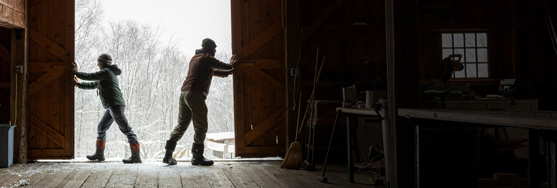 Two people pushing barn doors open with snow covered trees outside in the background