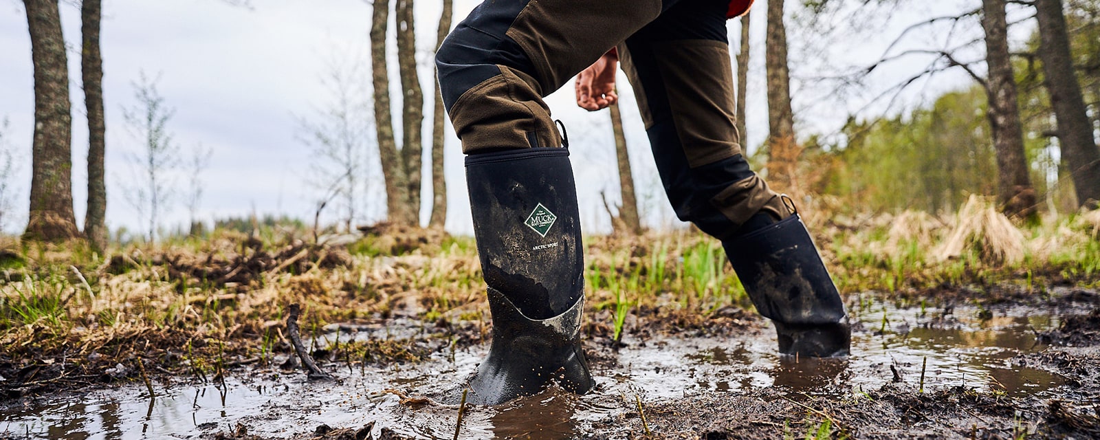 Close-up image of a man walking through a muddy woodland, wearing a pair of Muck Boots Arctic Sport wellingtons