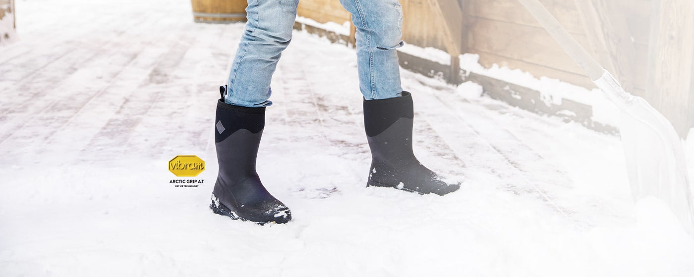 Person shovelling snow, wearing a pair of Muck Boots Arctic Ice Vibram wellingtons with a Vibram logo to the left of the image