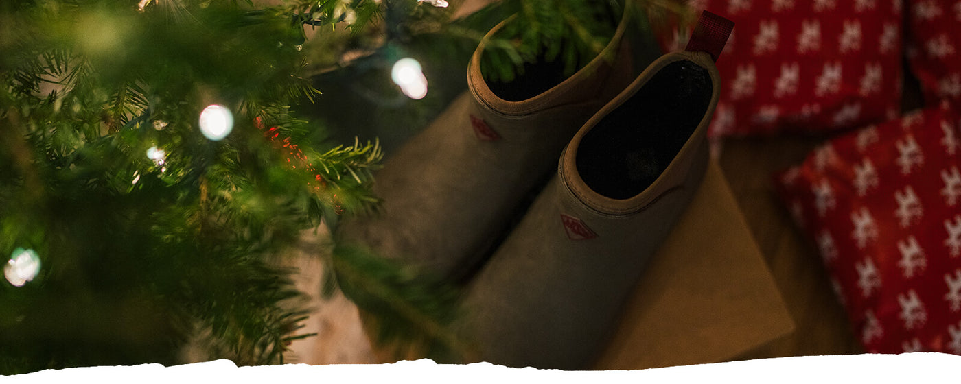 A pair of Muck Boots amongst a Christmas tree with lights and wrapped presents.