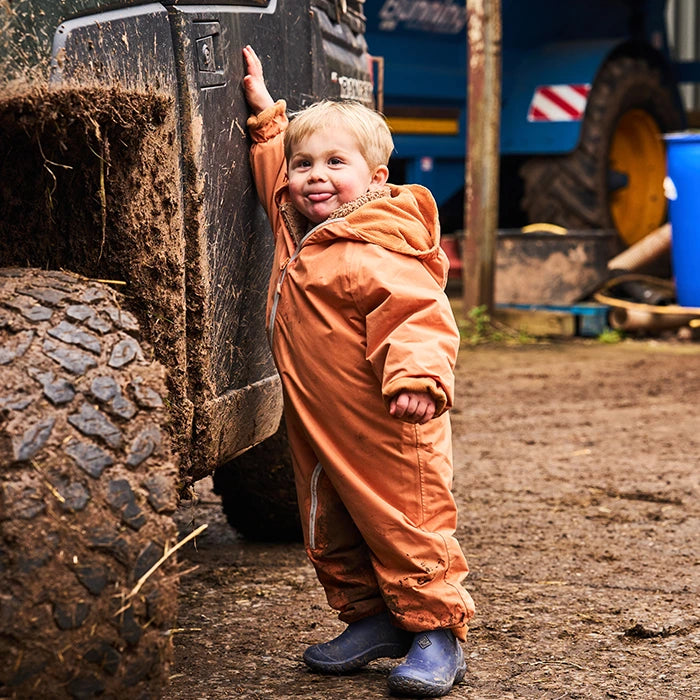 A little boy in an orange suit wearing a pair of blue Muck Boots and touching a muddy truck.