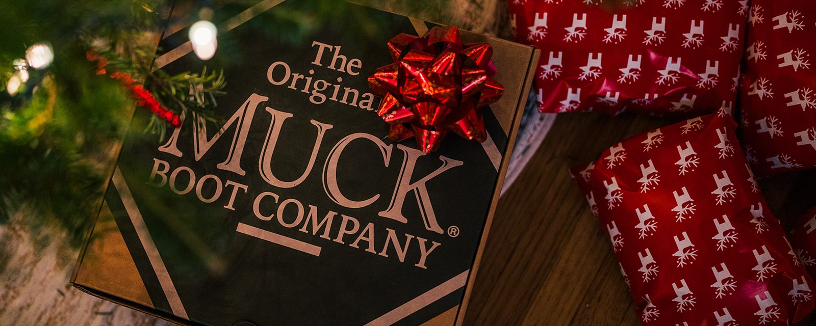 A Muck Boots footwear box with a Christmas gift bow under a Christmas tree and wrapped presents