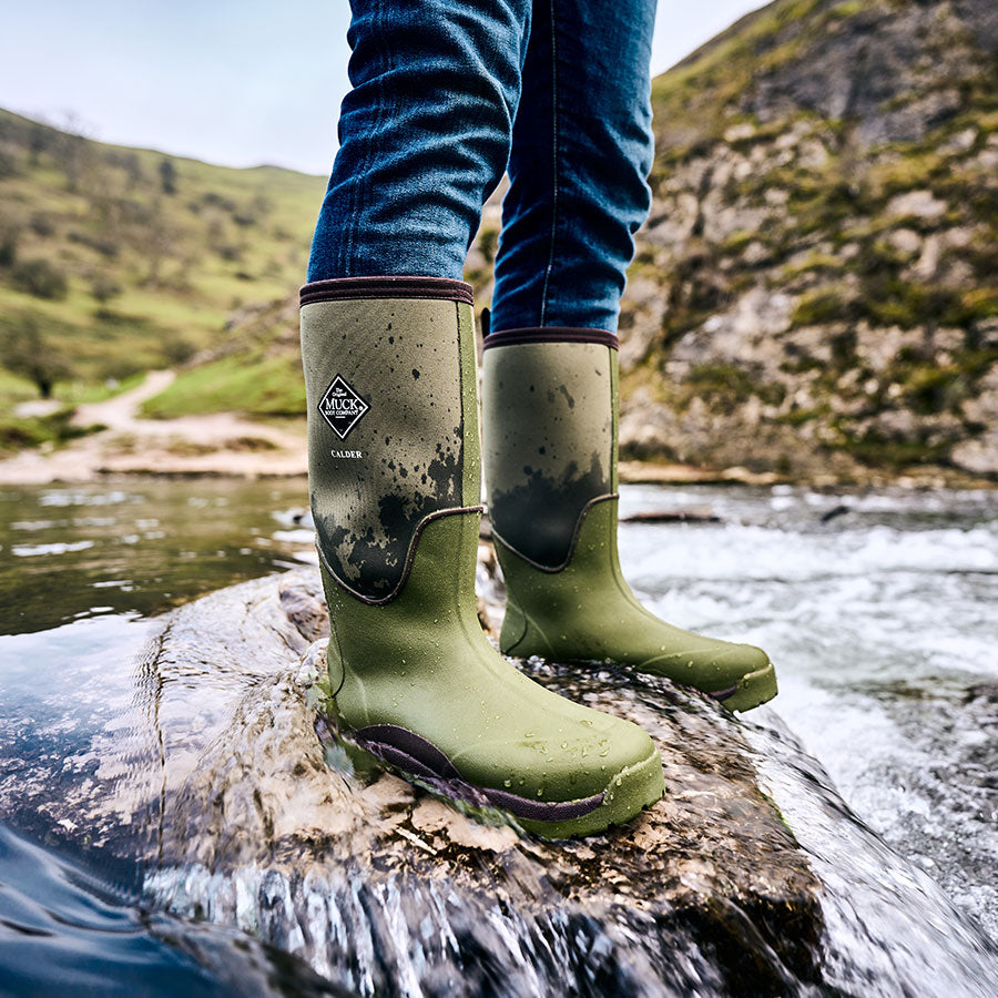 Close up of a man wearing a pair of green Muck Boot Calder Wellingtons, stood on a rock in a fast-flowing river