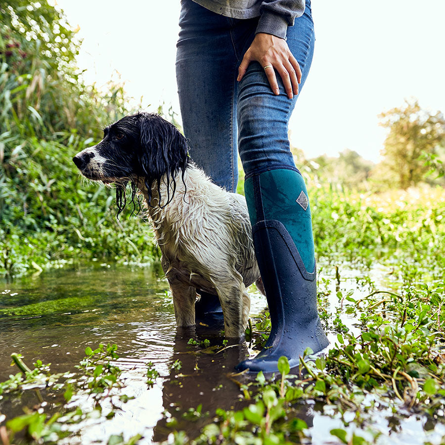 Close up image of a woman wearing a pair of Muck Boot Arctic Sport II Tall Boots with a wet black and white dog, stood in a large puddle with vegetation around them