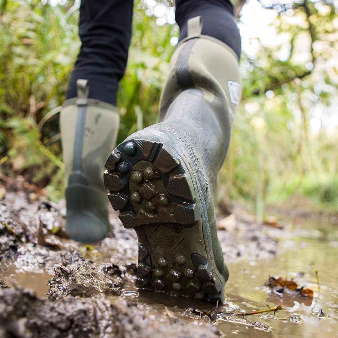 Close-up image of a person walking along a muddy path, wearing a pair of Muck Boots Derwent wellingtons with an outsole showing