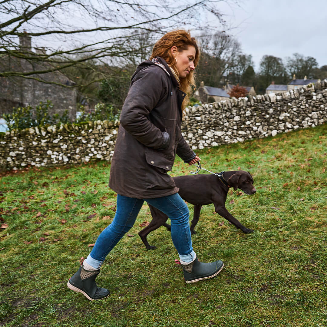 Woman wearing a pair of Muck Boots walking a black dog on a grassy field with trees and a stone wall in the background