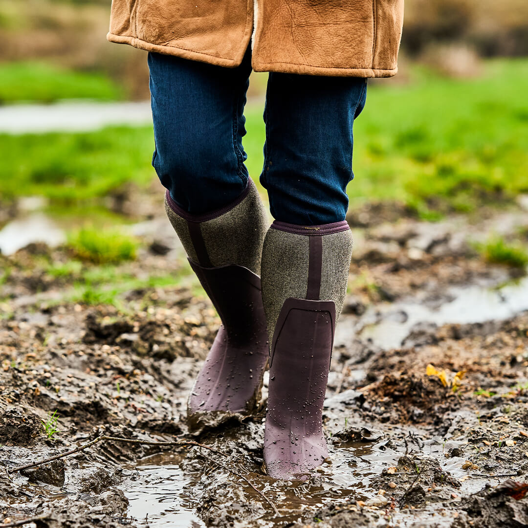Person wearing a pair of Muck Boots Arctic Sport Tall wellingtons and a coat walking through a muddy field.