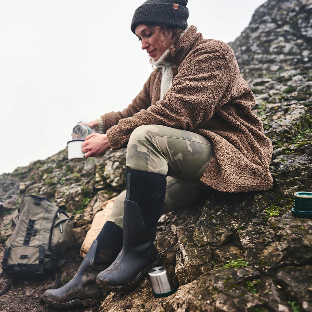 Woman sat on a rocky hill, pouring a hot drink into a cup and wearing a pair of Muck Boots wellingtons.